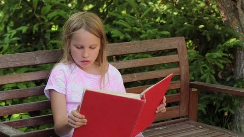Child reading book aloud while sitting on a wooden bench in a park Stock Footage 304149290