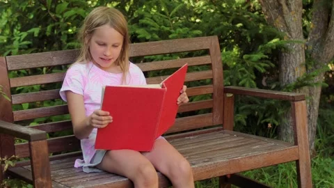 Child reading book aloud while sitting on a wooden bench in a park Stock Footage 304149378