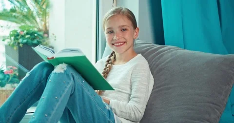 Child reading book and looking at window in rainy day and smiling at camera Stock Footage 77284179