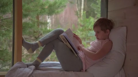 The child is reading a book. The boy settled down comfortably on the windowsill Stock Footage 165553472