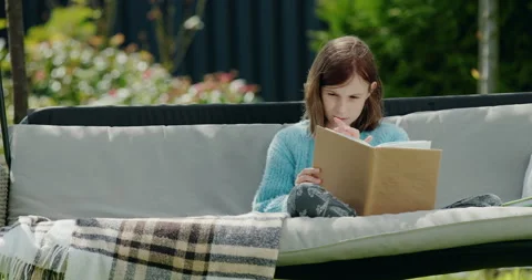 Child reading a book, sitting in a garden swing in the backyard of a house Stock Footage 249963833