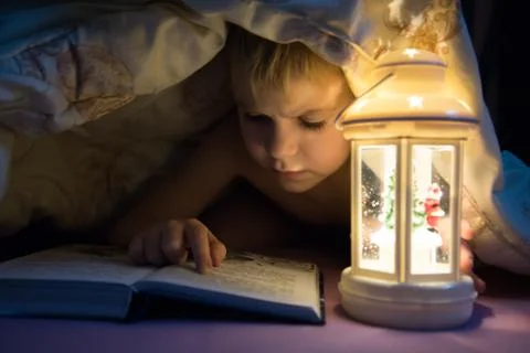 A child is reading a book under the covers. A child lights a book using a New Stock Photos