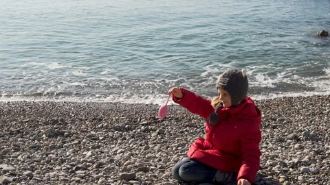 A child in a red jacket and hat plays on the rocky shore of the sea in the cold  Video stock 121313012