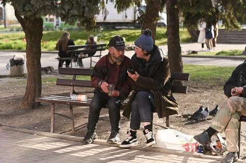 Child rides bicycle in park while homeless people sit on bench. Фото
