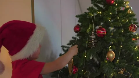 Child in Santa's red hat decorates Christmas tree at home evening indoors Stock Footage 220917355