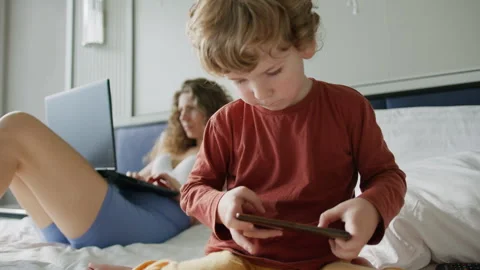 Child sits on a bed, absorbed in a mobile device, while an adult works intently Stock Footage 296512061