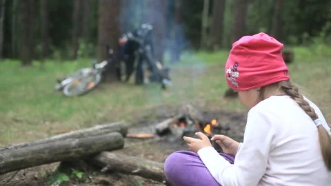 Child sits by fire and play smartphone. girl watching phone. Bicycles, bonfire Stock Footage 117290107