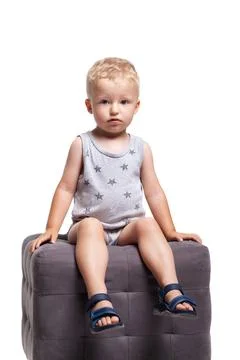 Child sits on a gray cube wearing a light outfit and sandals in a bright indoor Stock Photos