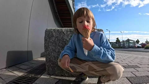Child Sits by a Rock and Eats Strawberry-Flavored Red Ice Cream Stock Footage 253871434