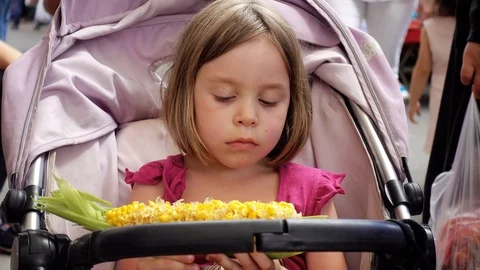 Child sits in a stroller and eats a boar of boiled corn. Girl 5-6 years of Eu Stock Footage 92347125