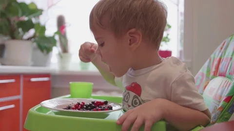 The child sits at the table and eats a spoonful of fresh berries. Useful and Vidéo 82652302