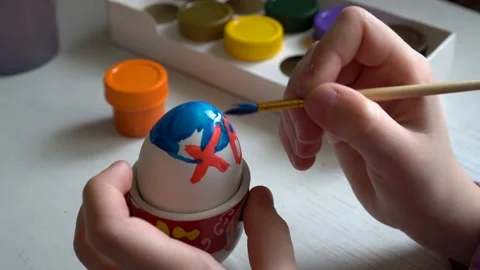 The child sits at the table and paints a brush Easter egg blue and red paint Видео 103166090