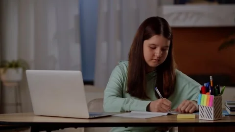 The child sits at a table in front of a computer and makes notes in a notebook Stock Footage 241924303