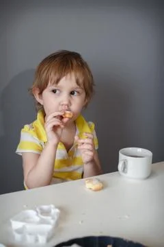 The child sits at a white table by the gray wall, drinking tea and biting coo Foto stock
