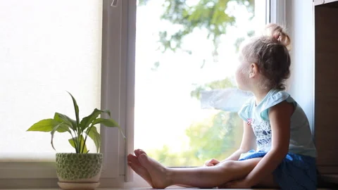 A child sits on a window sill and looks out the window. Stock Footage 92887772
