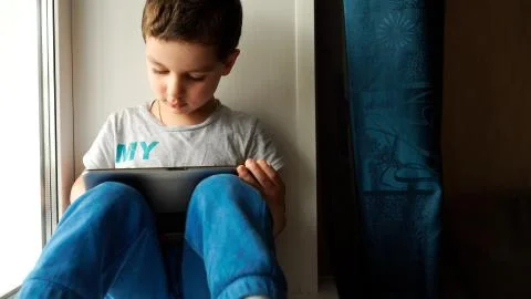 Child sits on a windowsill and uses a digital tablet pc Stock Photos