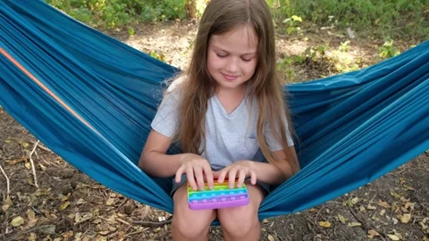 Child sitting in a hammock playing with pop it sensory toy. Girl presses on Video stock 159215362