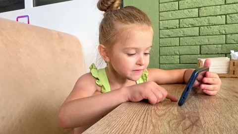 A child is sitting at a table in a cafe and playing games on the phone. Stock Footage 266163499
