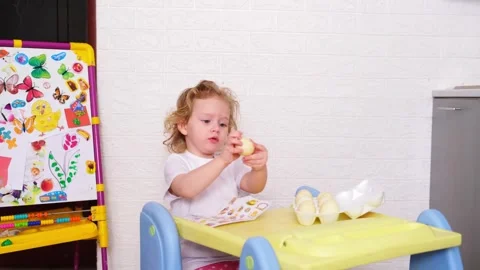 Child sitting at table ready to decorate Easter eggs, toddler holding an eg.. 動画素材 330109024