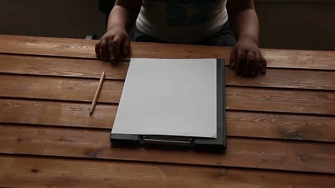 The child is sitting at the table on which lies a clean white sheet and pencil Stock Footage 75559001