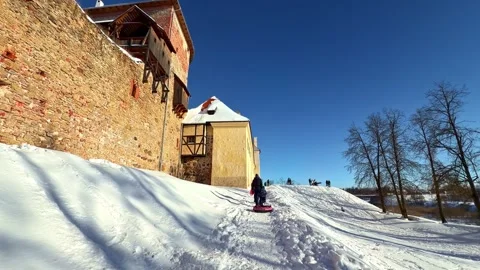 Child sledding down a snowy hill next to Bauska Castle in winter on a sunny day 스톡 동영상 342934901
