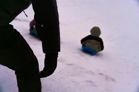 Child sledding on a snowy slope with a blurred perspective of an adult watching Stock Photos