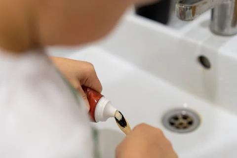 Child squeeze tooth paste onto bamboo brush over white sink in bathroom Fotos de archivo