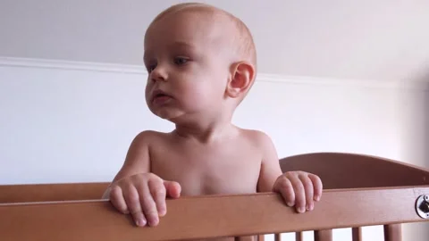 A child standing in a crib without a shirt holds on to the handrails and sadly Stock Footage 269671413