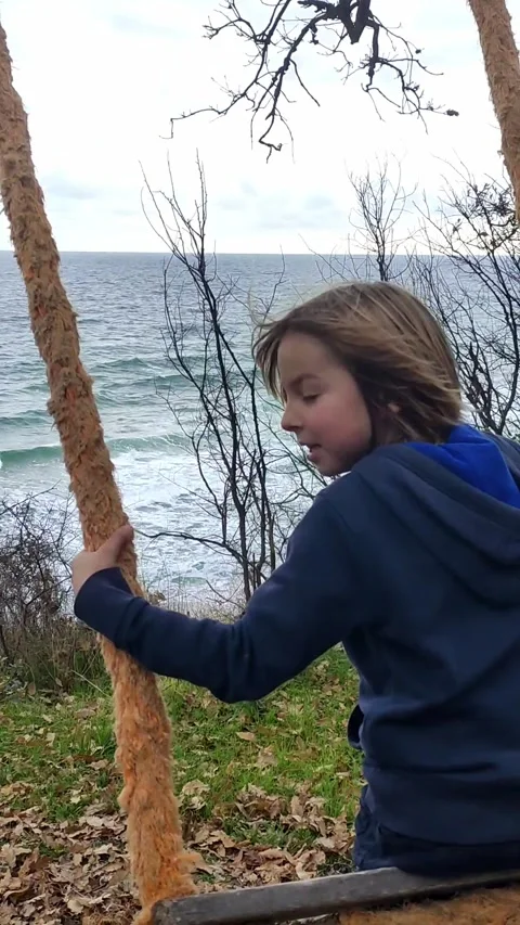 A child swinging on a rustic rope swing with an ocean view. The peaceful Stock Footage 298042847