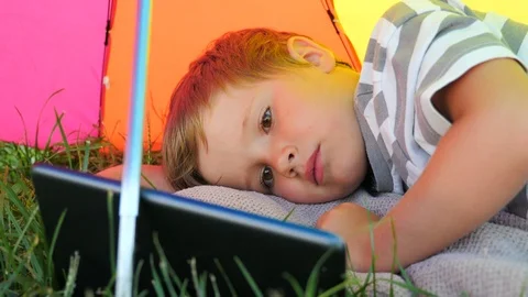 Child with tablet lying on green grass under sun umbrella. Summer vacation Stock Footage 97808675