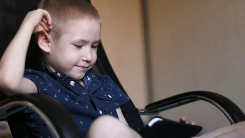 A child with a tablet, watching cartoons, smiling. Sits in an office chair Stock Footage 74979379