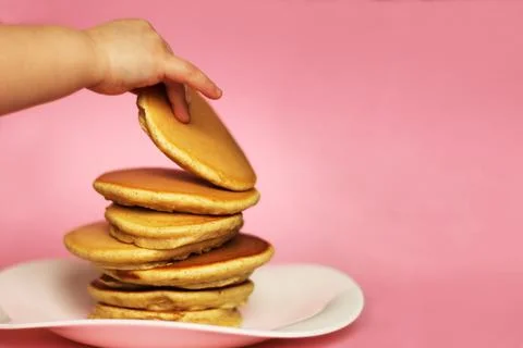 A child takes a pancake from a stack, pink background. Stock Photos