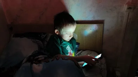 Child uses smartphone while sitting in bed at night in a dimly lit room Vídeo Stock 336252310
