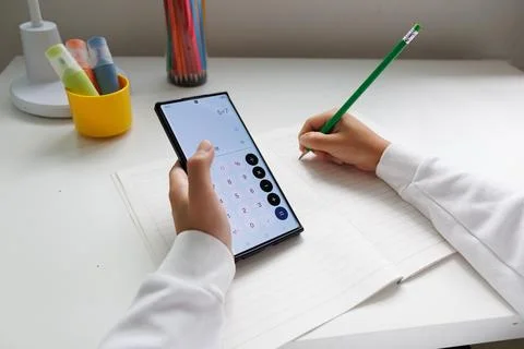 Child using a smartphone calculator while writing answers in a notebook durin Stock Photos