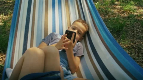 Child using a smartphone while relaxing in a striped hammock outdoors. Digital Stock Footage 264339129