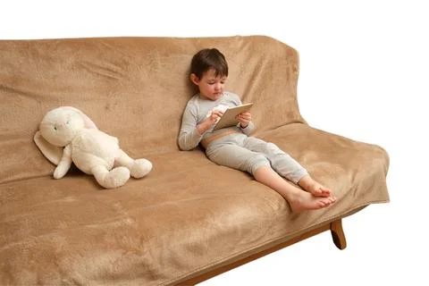 Child using tablet while sitting on a brown sofa with plush rabbit toy. Kid r Stock Photos