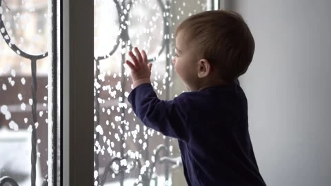 The child watches the snowfall through the window from the house. A little son Stock Footage 231402697