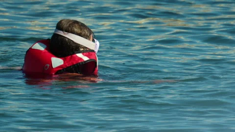 Child wearing a red life jacket and goggles while swimming in calm blue water, Stock Footage 339802281