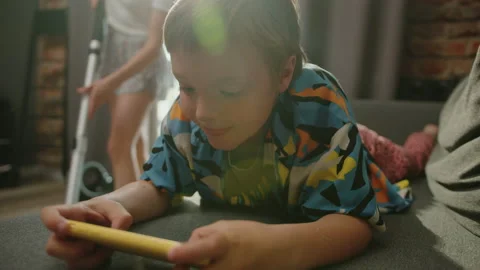 Child who is playing on the phone while his mom is vacuuming in the room Stock Footage 233777817