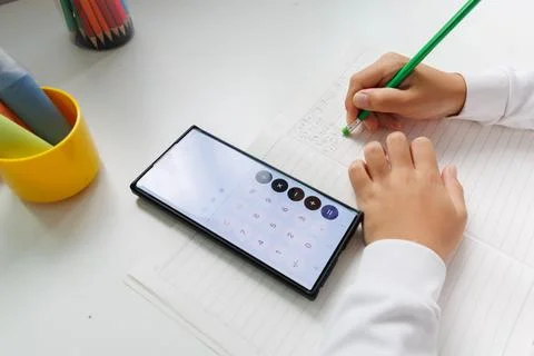 Child writing math answers in a notebook after calculating with a smartphone. Stock Photos