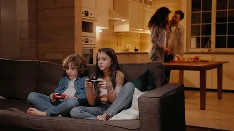 Children are playing on game console, parents preparing dinner at the background Видео 165412575