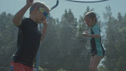 Children bouncing high on trampoline playing in summer with water Video stock 135797436