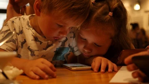 Children at cafe table enjoy watch mobile cartoons boy and girl bend over phone Stock Footage 78217424