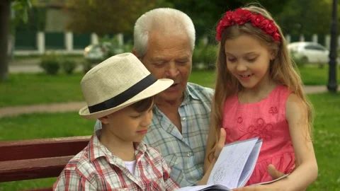 Children joyfully explore a book with their grandpa in a sunny park Stock Footage 311982049