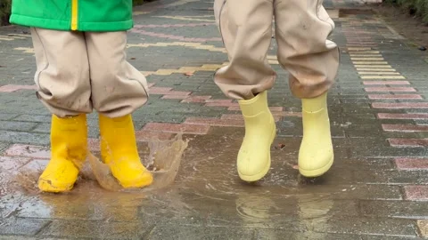 Children jumping in puddle wearing bright yellow rubber boots, puddle splashes Stock Footage 328352201