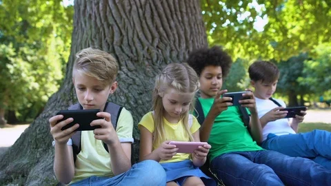 Children playing gadgets, sitting under tree in park, boy smiling in camera, app Vídeo Stock 117209192