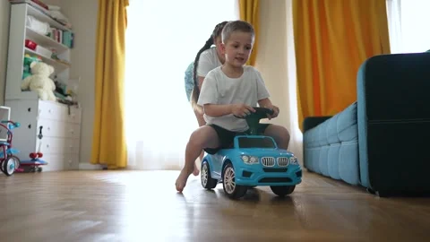 Children playing at home. Young boy rides toy car with sister in living room Stock Footage 345335980