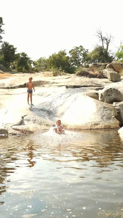 Children playing to jump down a stone slide into a natural pool in summer Stock Footage 277454016