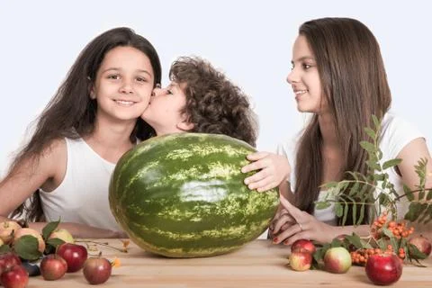 Children sit at the table in front of a large watermelon whole. Boy kisses gi Stock Photos