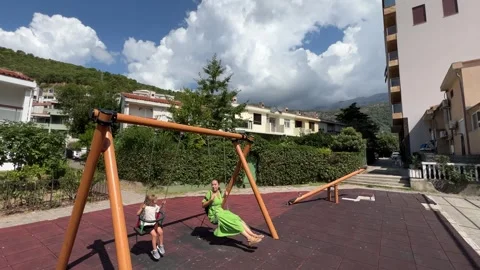 Children swinging under the cloudy sky in the park near the city buildings Vídeo Stock 266901113